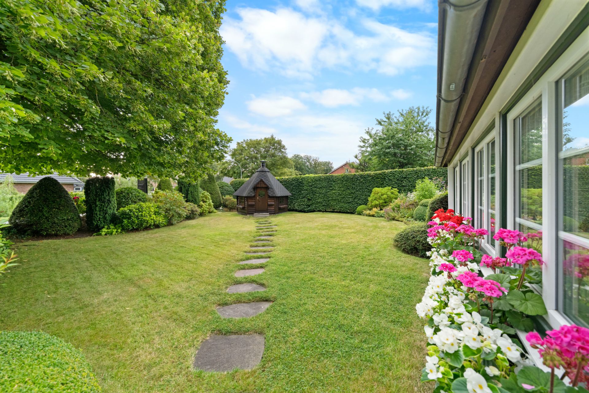 Grüner Garten mit Blumen, Steinen als Weg und kleinem Holzhaus im Hintergrund.