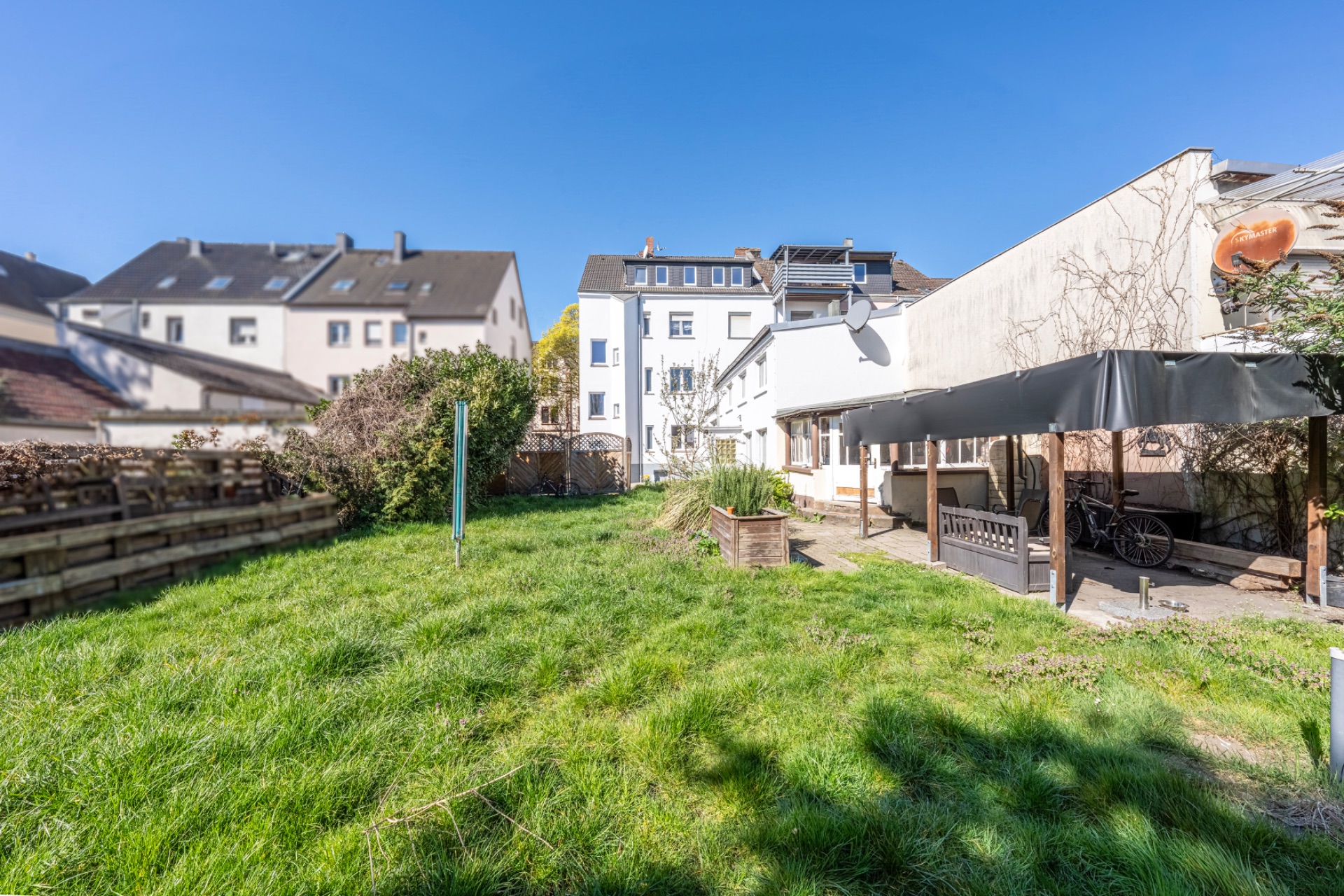 Grüner Garten mit überdachter Terrasse und Wohnhäusern im Hintergrund.