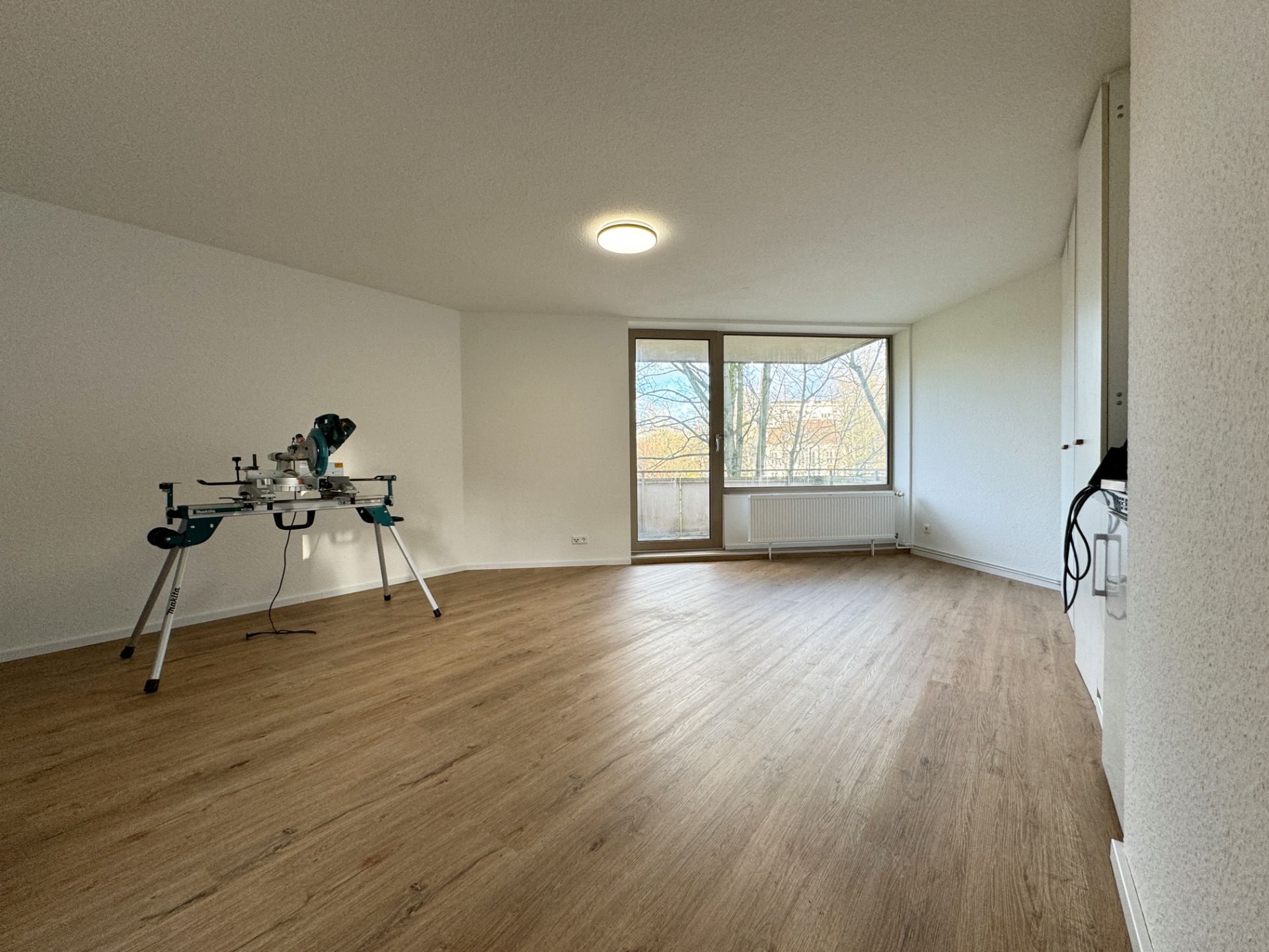 Helles Zimmer mit Holzfußboden, großem Fenster und einer Werkbank mit Säge links.