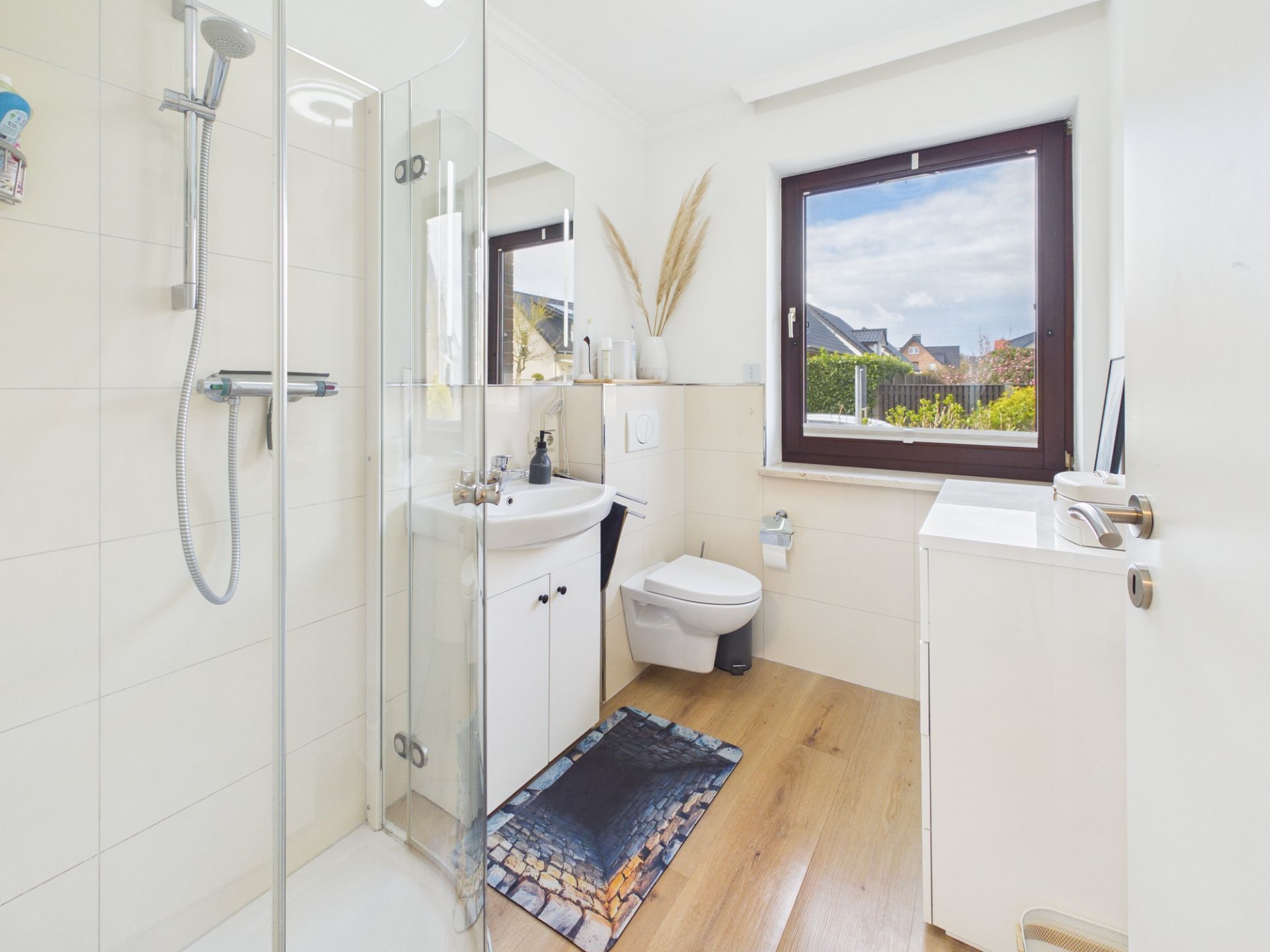 Helles Badezimmer mit Glasdusche, Waschbecken, WC, Fenster und Holzboden.