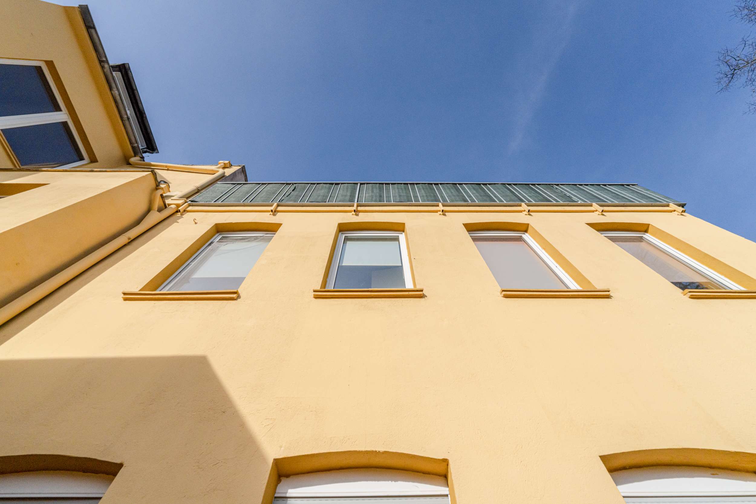 Gelbe Hausfassade mit mehreren Fenstern und Glasbalkon, fotografiert von unten bei blauem Himmel.