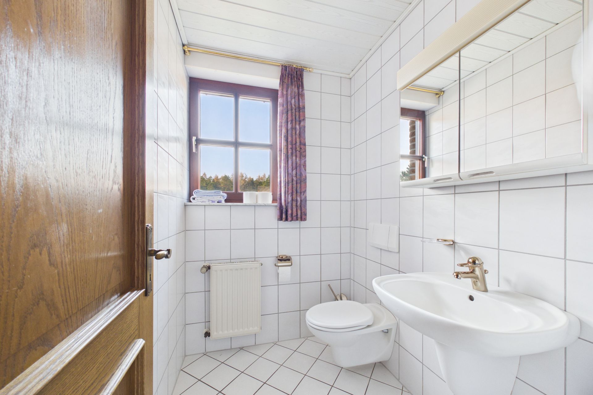 Helles Badezimmer mit weiß gefliesten Wänden, Waschbecken, WC und Fenster mit Vorhang.