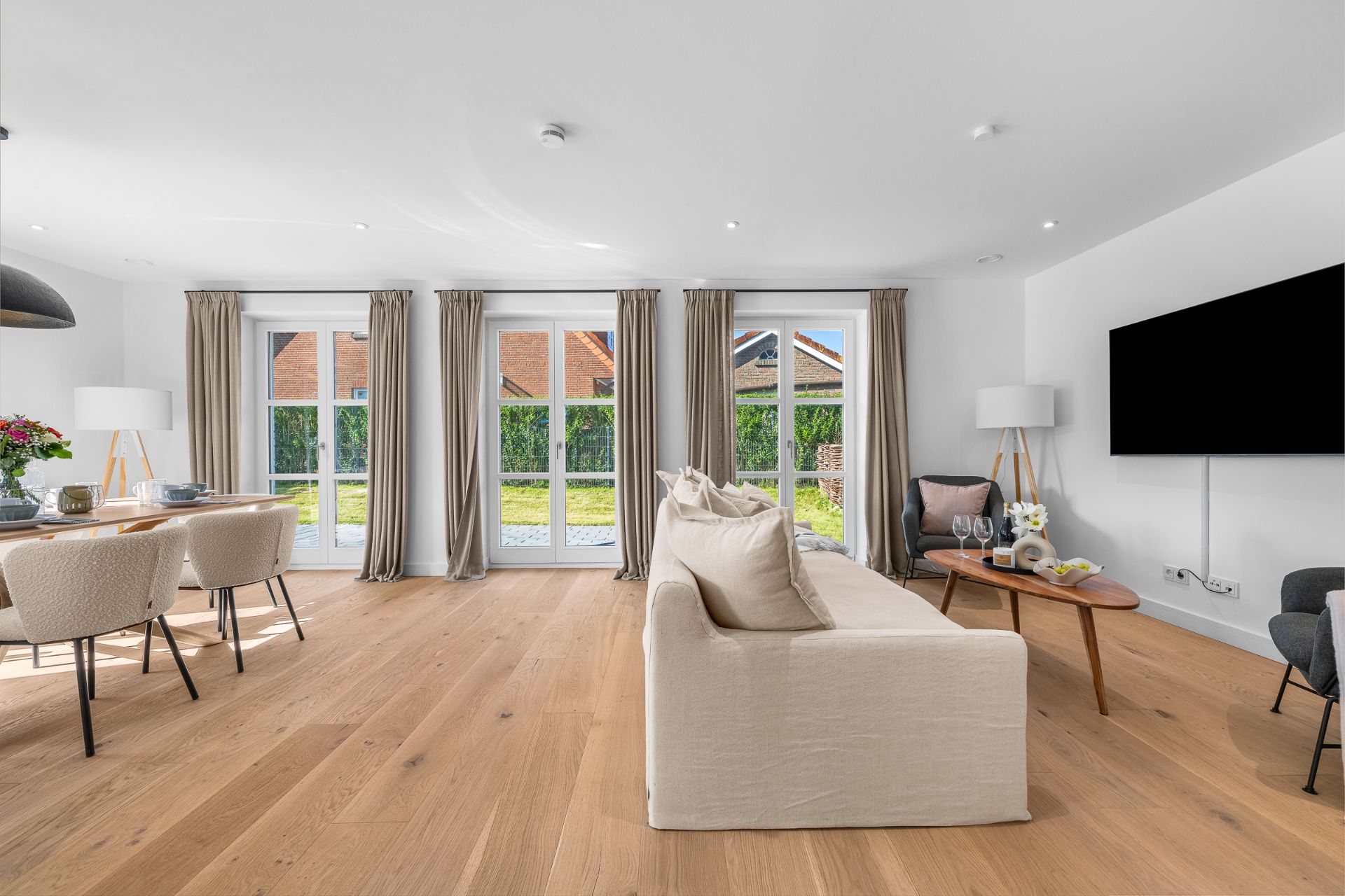 Helles Wohnzimmer mit Holzfußboden, Sofa, Esstisch, großen Fenstern und Fernseher an der Wand.