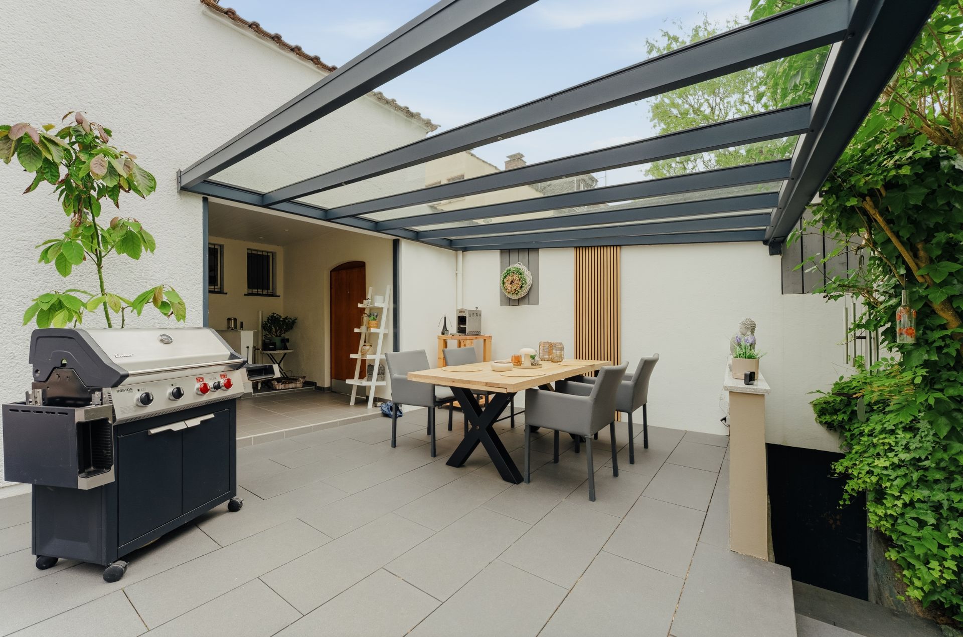 Überdachte Terrasse mit Esstisch, Stühlen und Gasgrill neben einer weiß gestrichenen Hauswand.