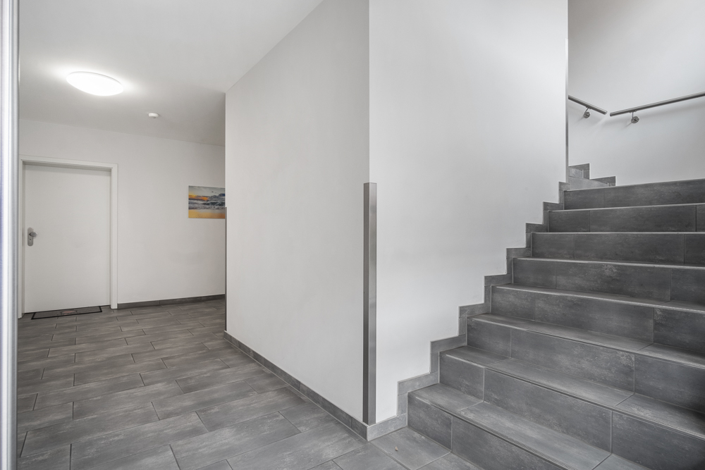 Heller Hausflur mit grauen Fliesen und Treppe, weiße Wände, Tür und Wandbild.