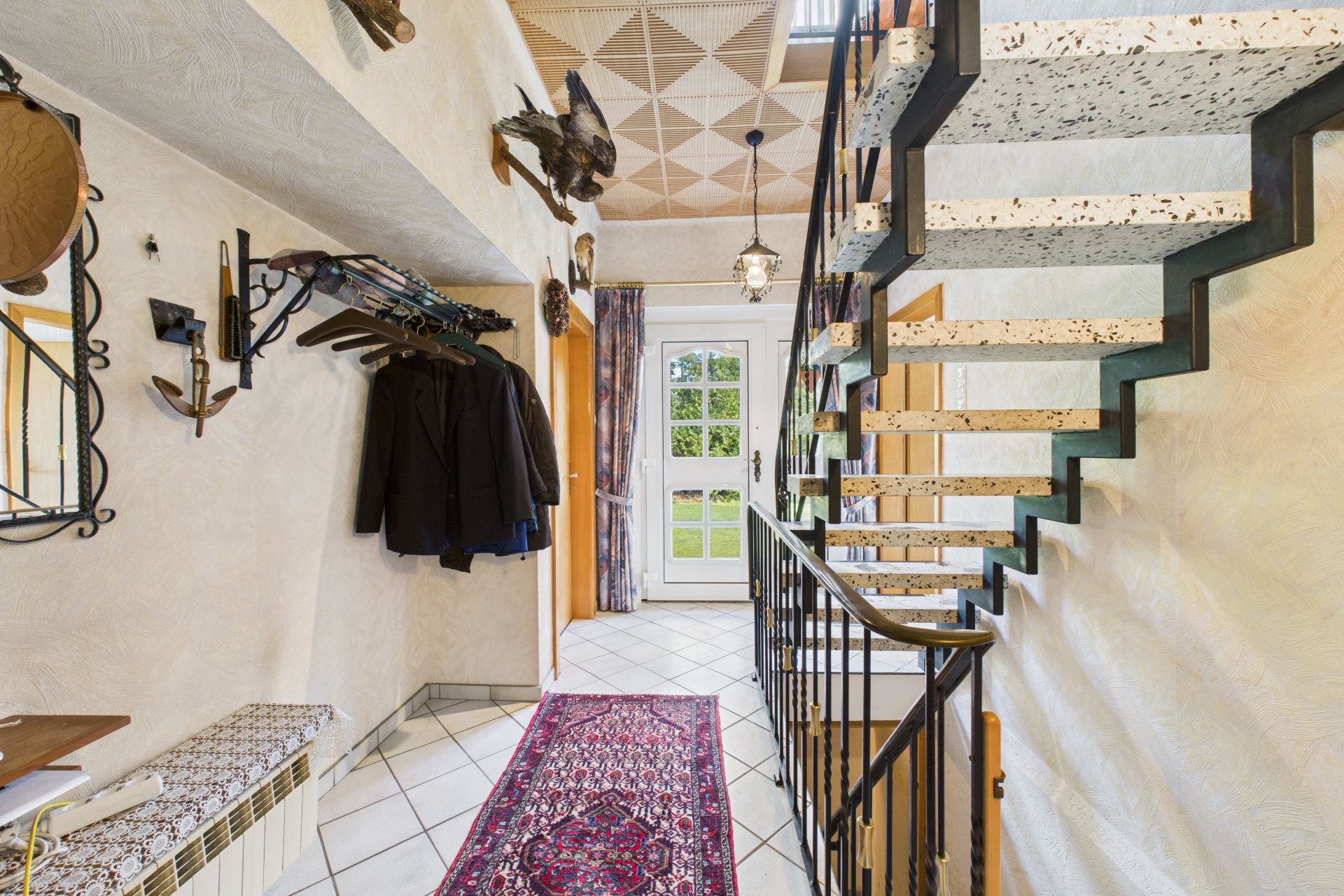 Flur mit Teppich, Garderobe mit Jacken, Treppe und Tür mit Blick auf den Garten.