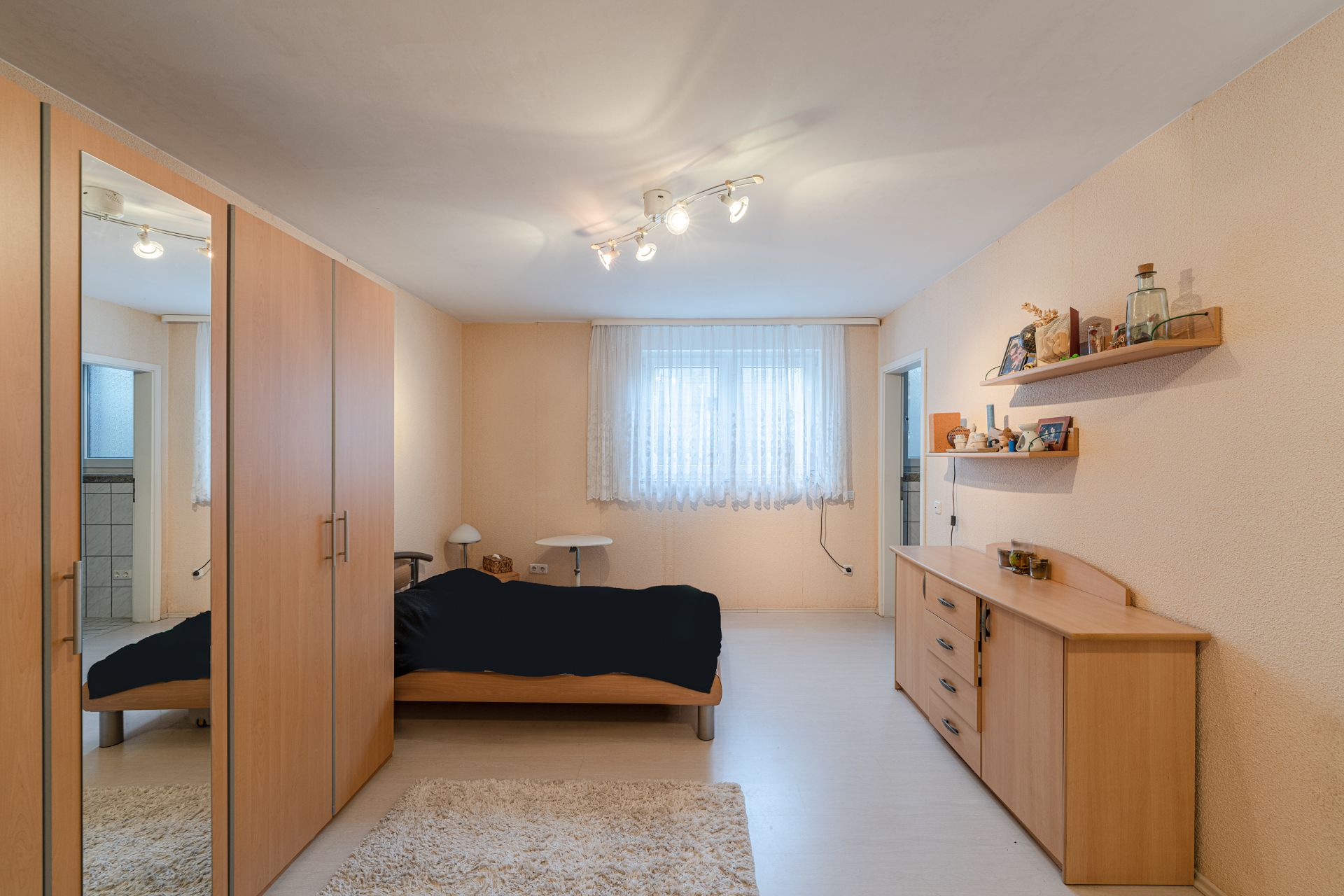Schlafzimmer mit Bett, großem Kleiderschrank, Kommode und Regal, Fenster mit Gardine.