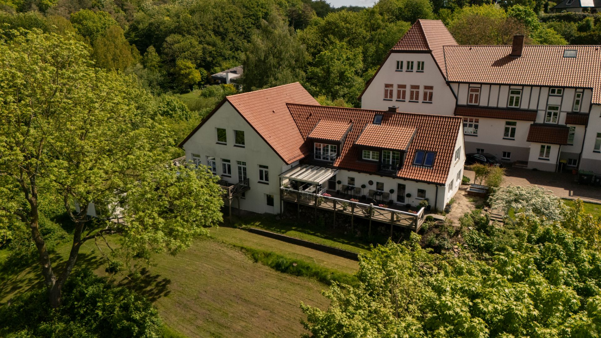 Großes Wohnhaus mit rotem Dach, Terrasse und umgeben von Bäumen und Grünflächen.