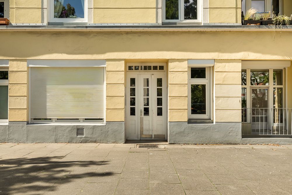 Gelbe Hausfassade mit weißer Tür, Fenstern und geschlossenen Rollläden, davor Bürgersteig.