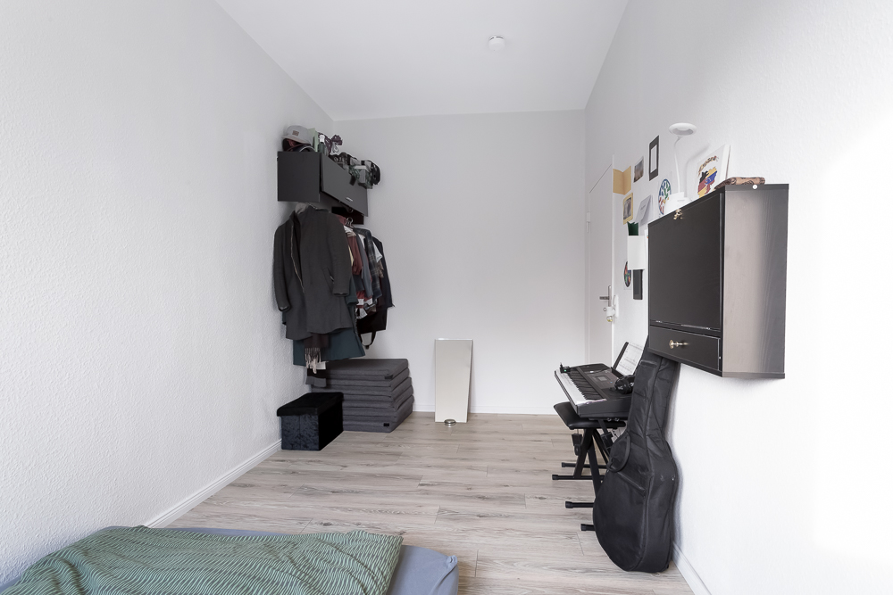 Helles Zimmer mit Kleiderstange, einem Keyboard, Gitarrentasche und Wandregal.