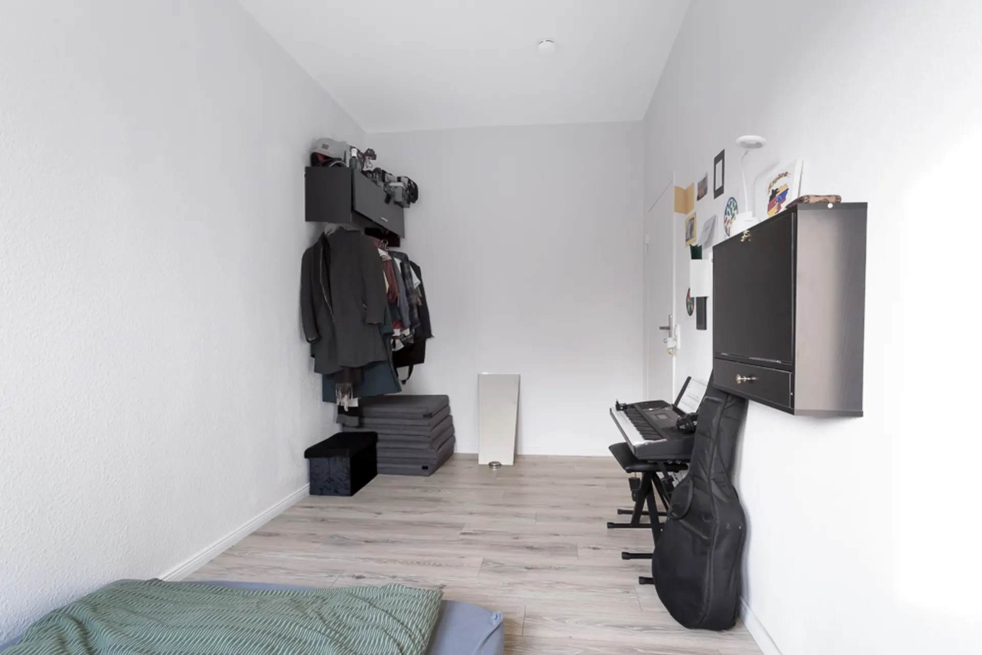 Helles Zimmer mit Kleiderstange, einem Keyboard, Gitarrentasche und Wandregal.