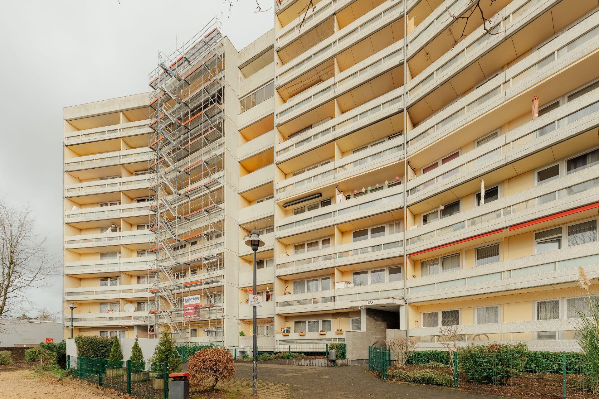 Großes Wohngebäude mit mehreren Balkonen, links Baugerüst, im Vordergrund einige Büsche.