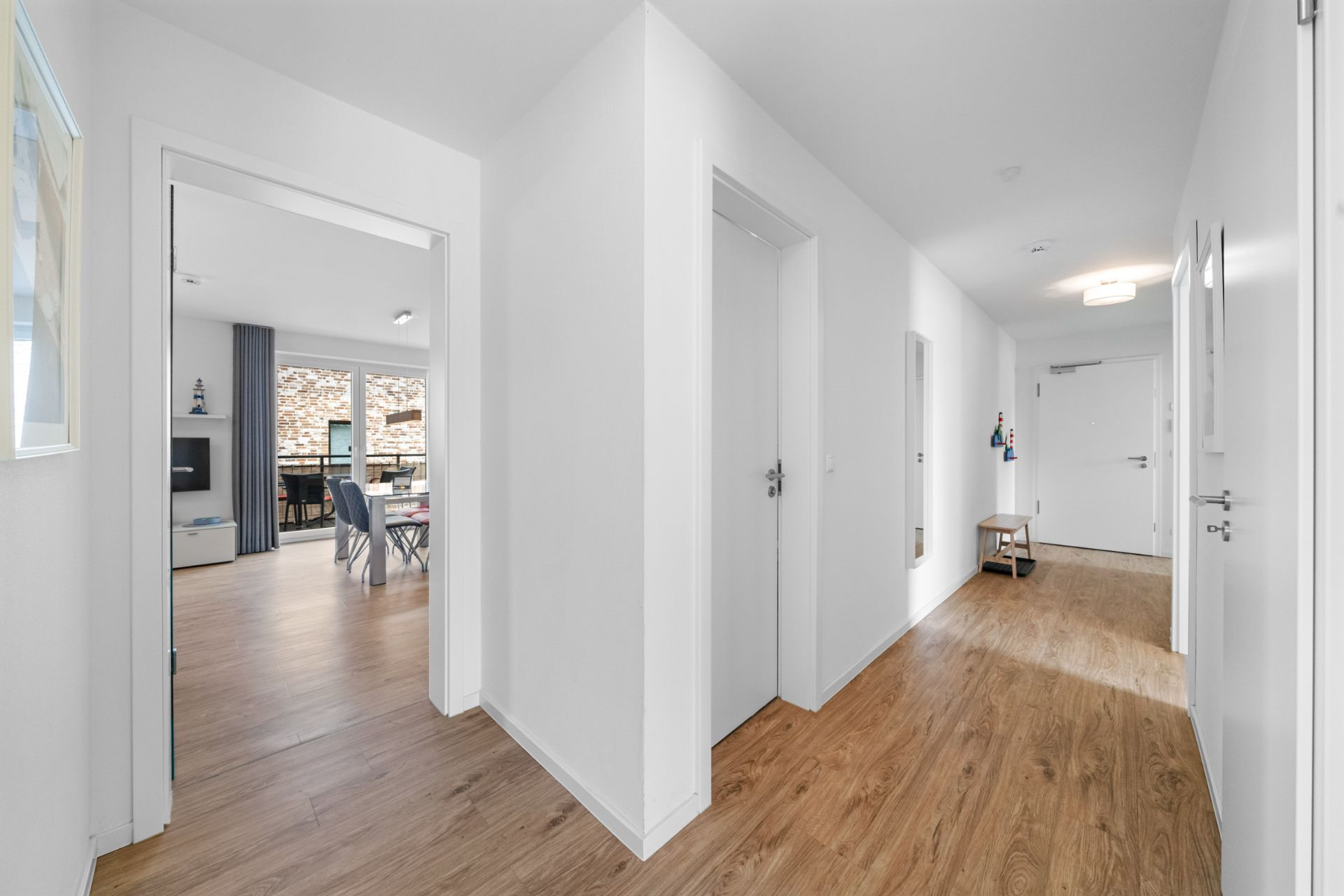 Heller Flur mit Holzboden, weißen Wänden und mehreren Türen, Blick in ein Wohnzimmer.