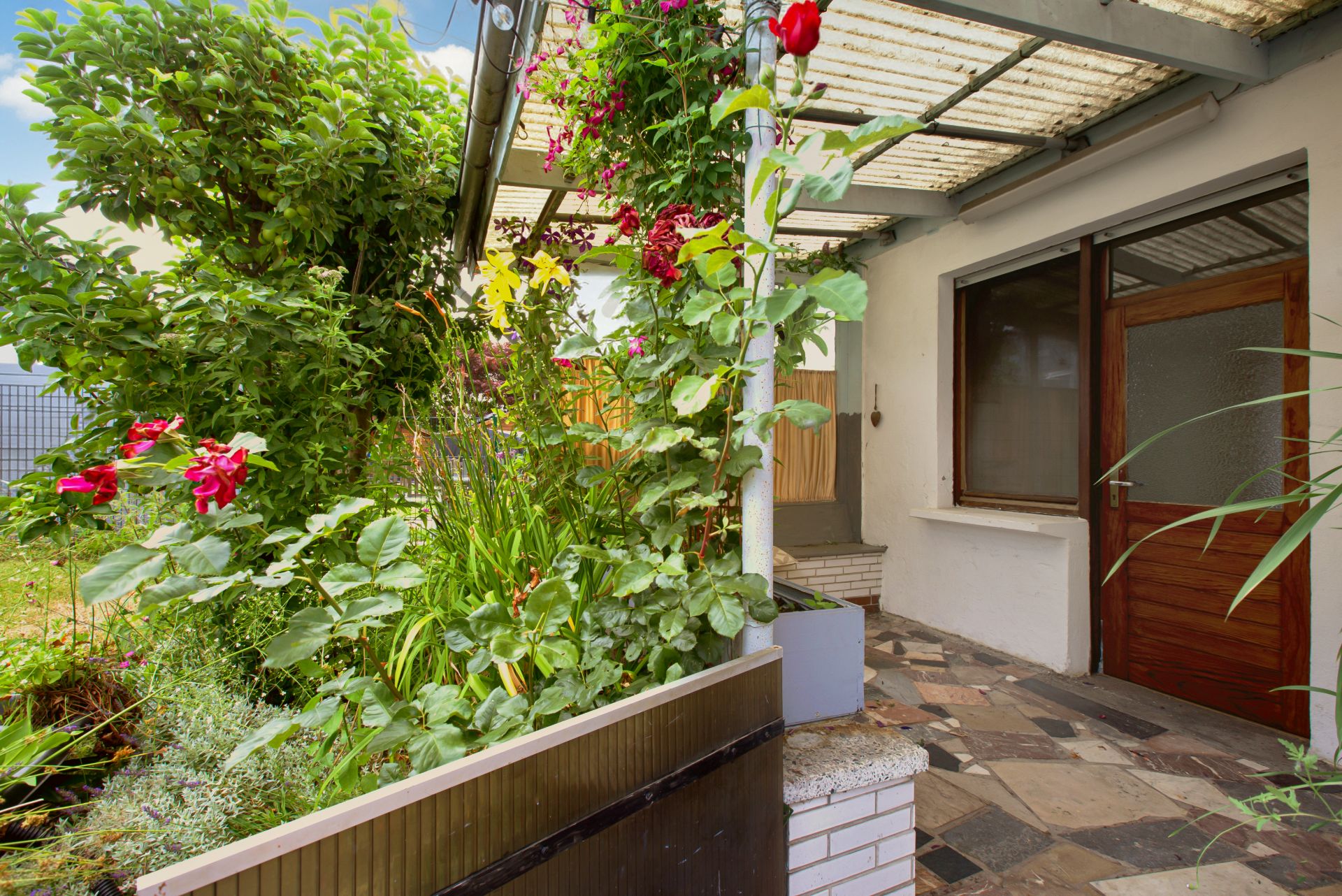Überdachte Terrasse mit Steinboden, Holztür und vielen grünen Pflanzen im Vorgarten.