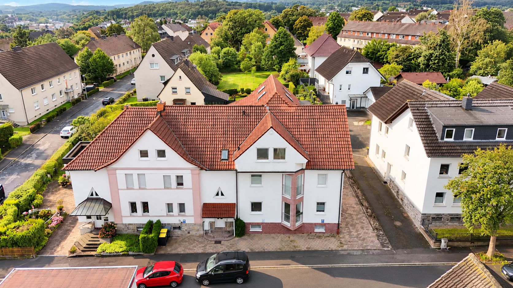 Mehrfamilienhaus mit rotem Dach, Vorgarten und geparkten Autos vor dem Gebäude.