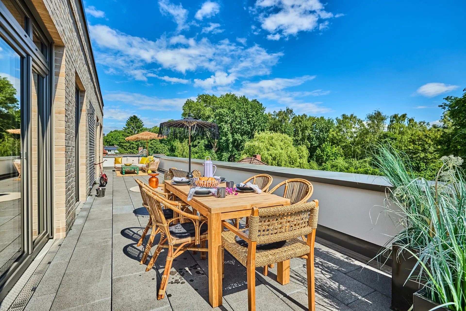 Terrasse mit Holztisch, Stühlen, Sonnenschirm und Blick auf Bäume unter blauem Himmel.