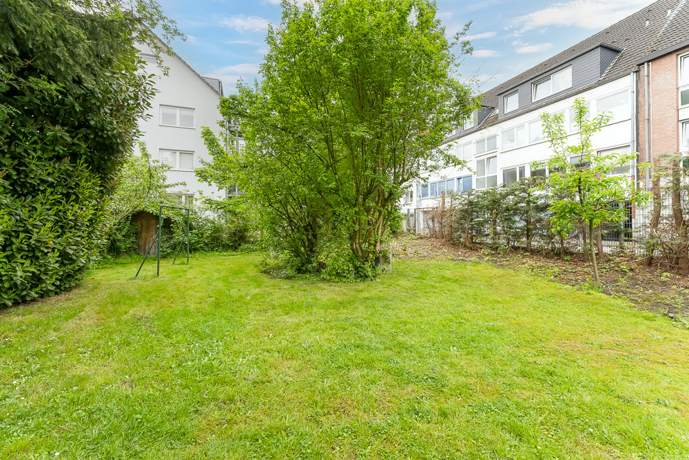 Grüner Garten mit Bäumen und Rasen, umgeben von Nachbarhäusern und Büschen.