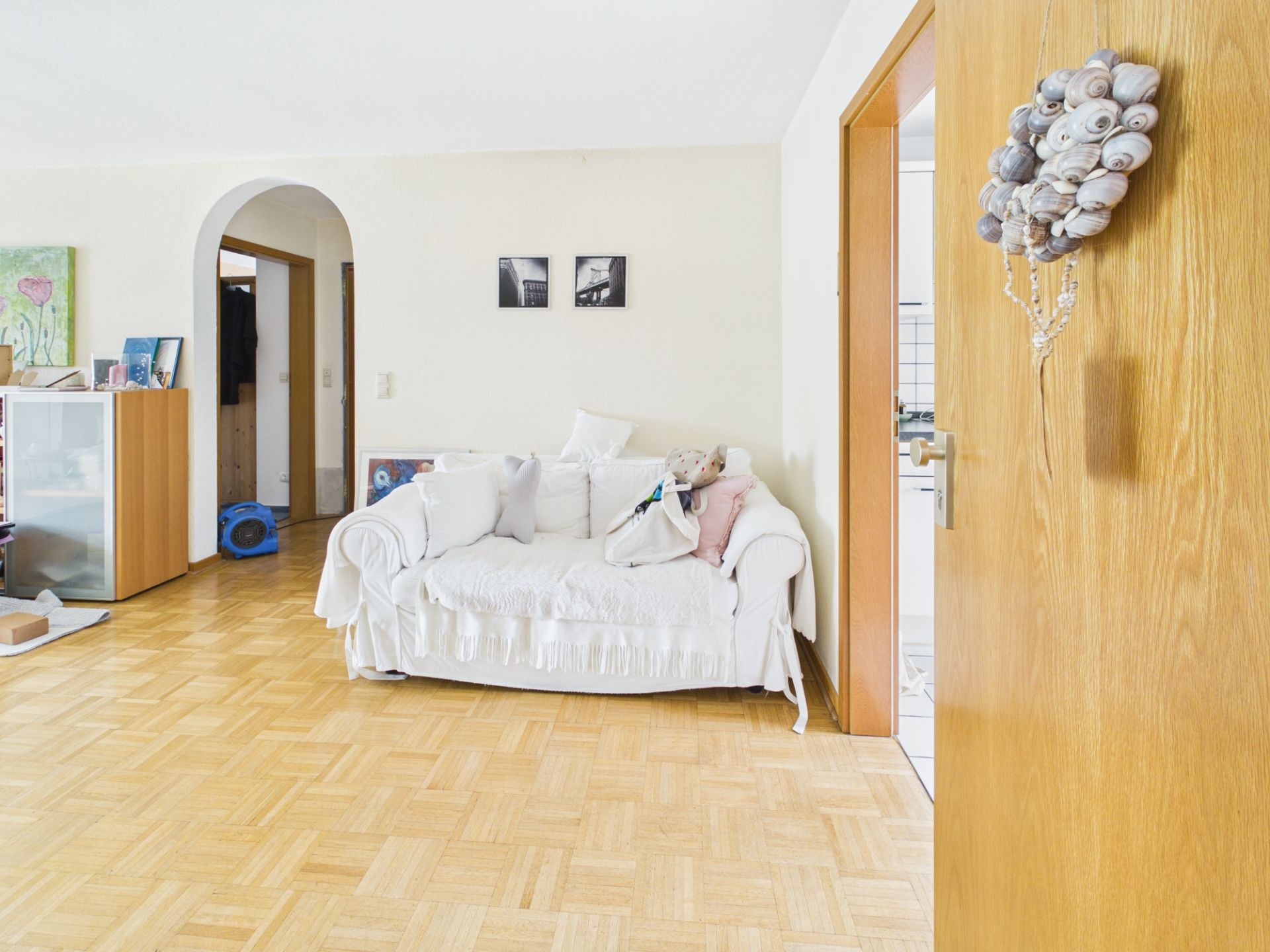 Helles Wohnzimmer mit weißem Sofa, Holzboden und offener Tür zu weiterer Räumlichkeit.