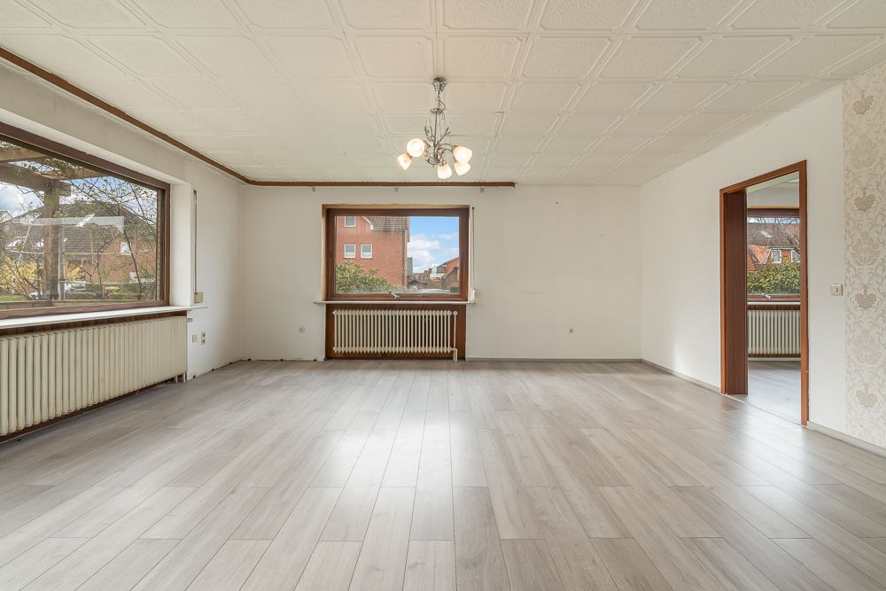 Helles, leeres Wohnzimmer mit großen Fenstern, Laminatboden und Deckenleuchte.