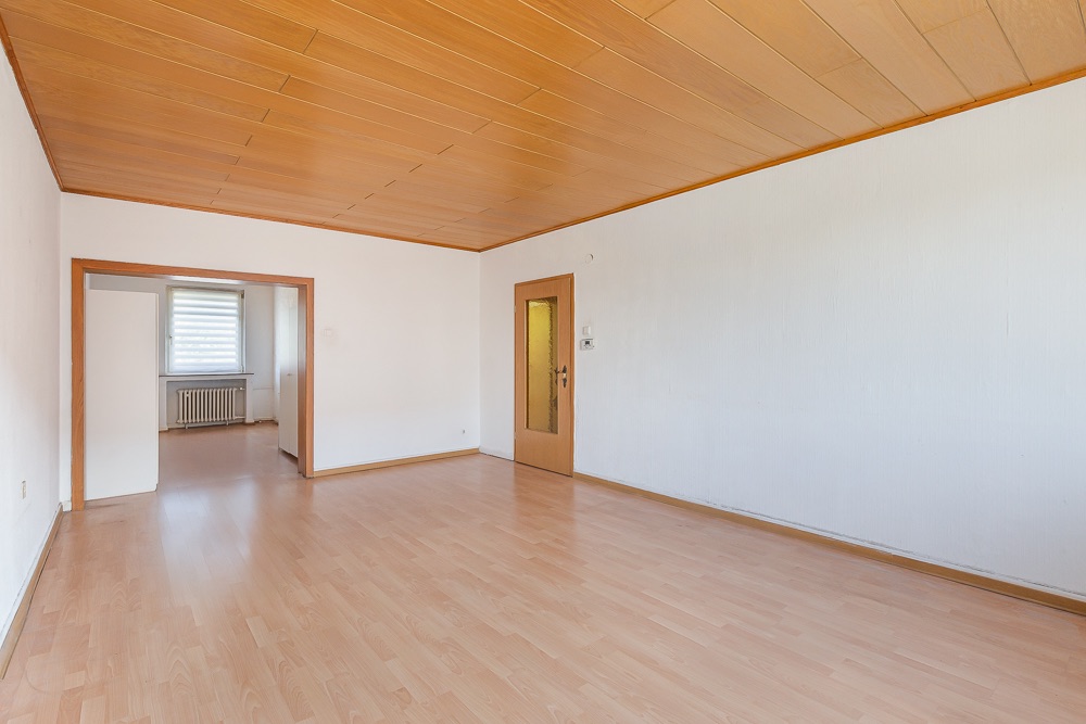 Leerer Raum mit Holzdecke, Holzboden und Tür; Durchgang zu weiterem Zimmer mit Fenster.