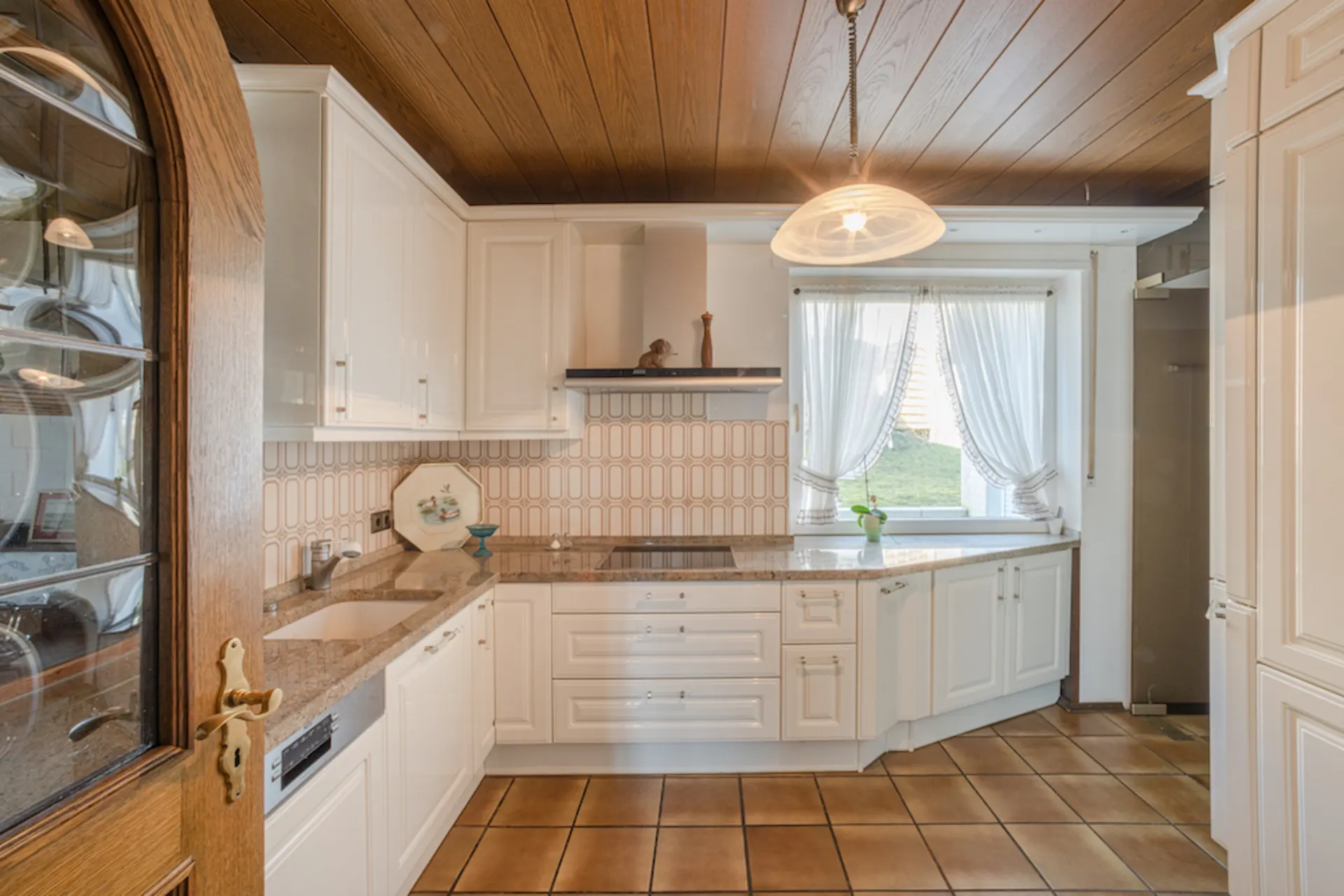 Helle Küche mit weißen Schränken, Holzdecke, Fliesenboden und Fenster mit Vorhängen.