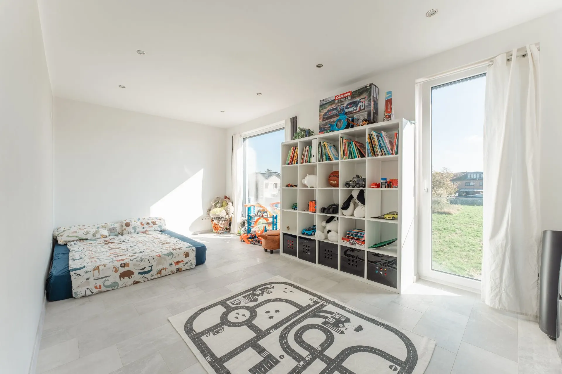 Helles Kinderzimmer mit Bett, Regal voller Spielzeug und Büchern, Spielteppich und großen Fenstern.