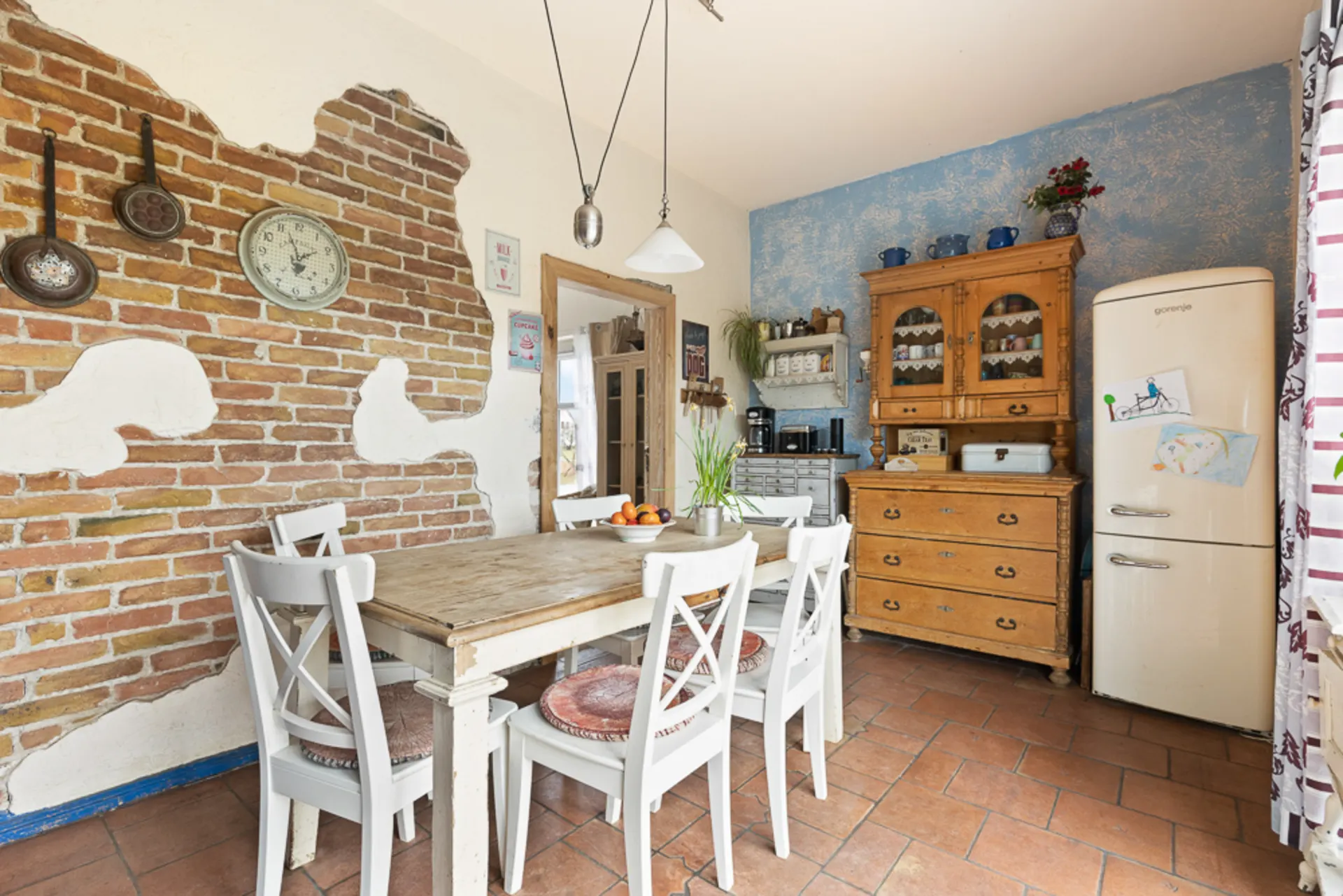 Gemütliche Küche mit Holztisch, weißen Stühlen, Backsteinwand, Vintage-Kühlschrank und Holzschrank.