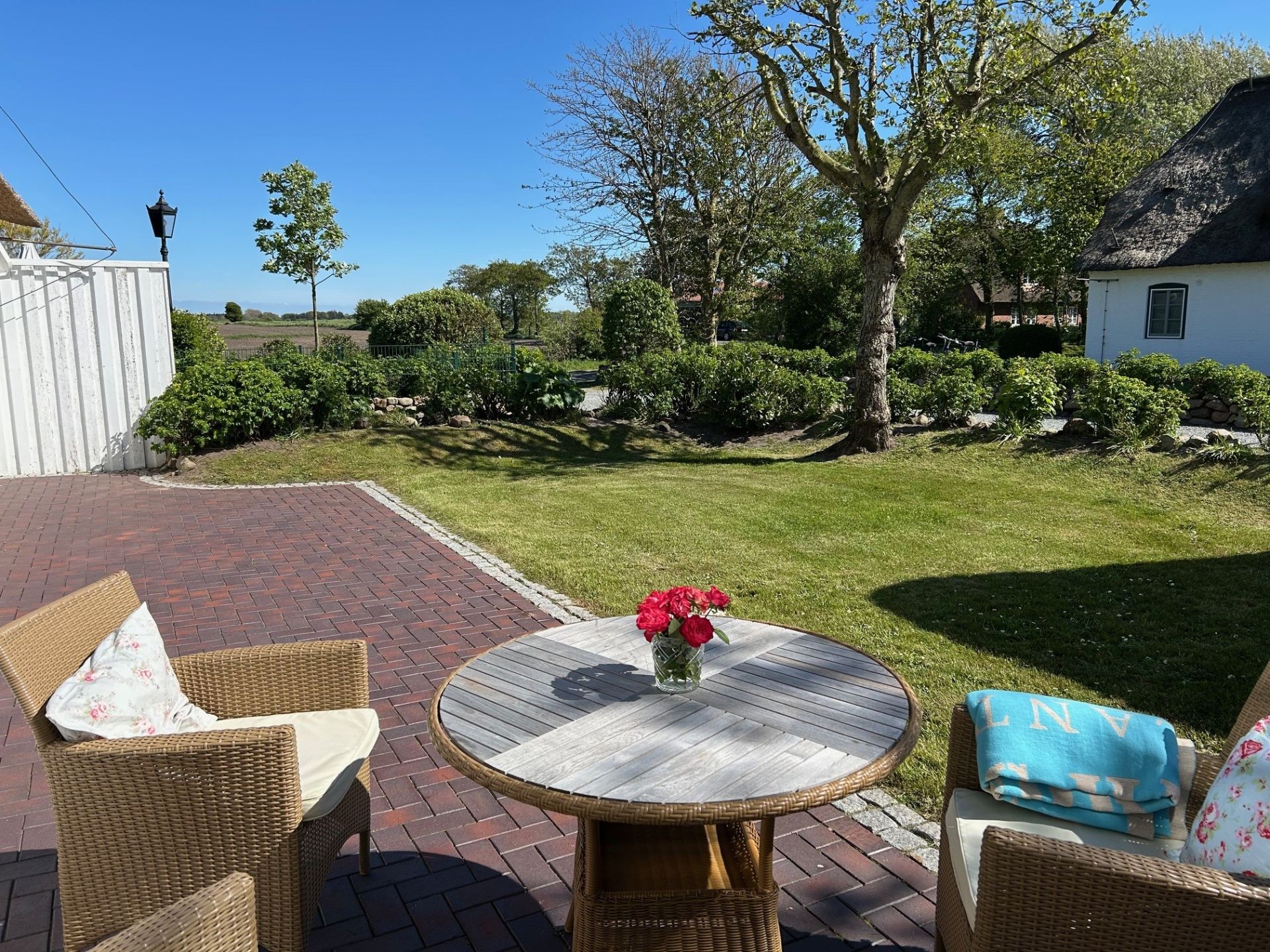 Terrasse mit rundem Holztisch, drei Korbstühlen und Vase mit Blumen, angrenzender Garten und Bäume.