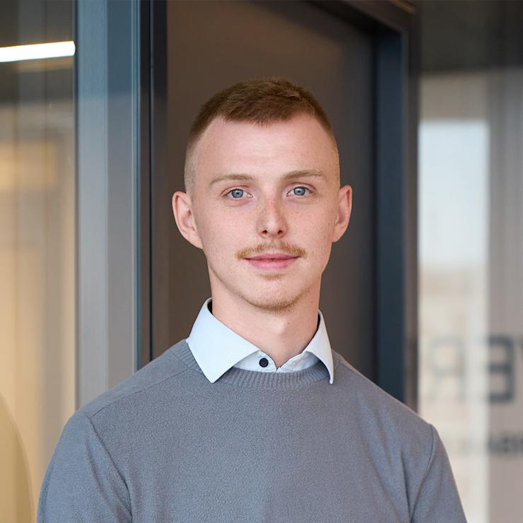 Junger Mann mit kurzem Haar und grauem Pullover steht vor einer Glastür.