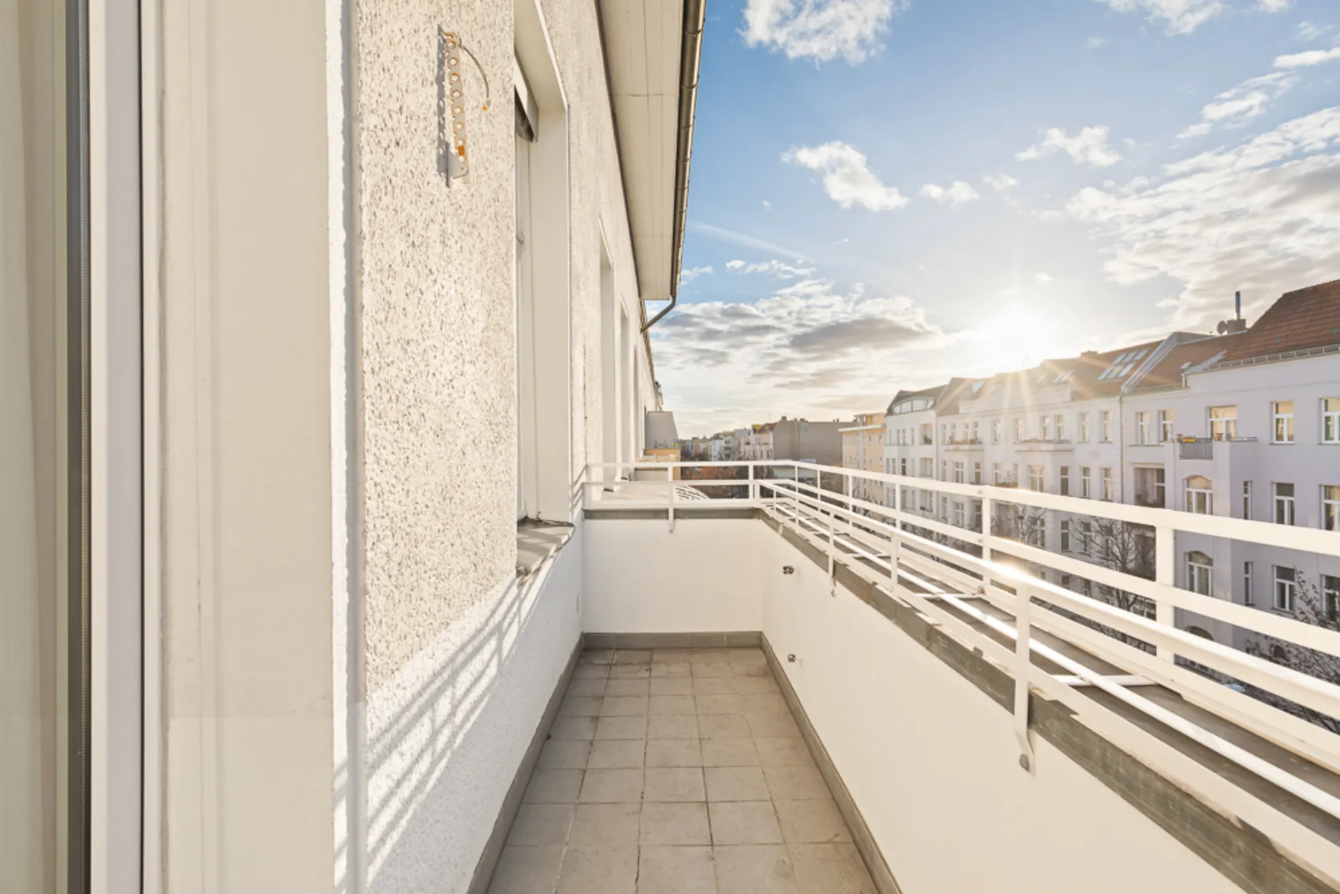 Heller Balkon mit Fliesenboden und weißem Geländer, Blick auf Nachbarhäuser bei Sonnenuntergang.