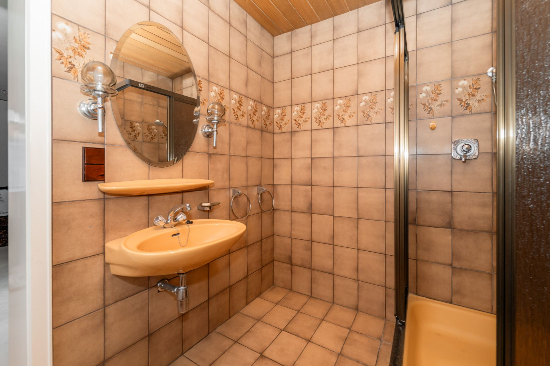 Badezimmer mit beigen Fliesen, Waschbecken, Spiegel, Ablage und Dusche.