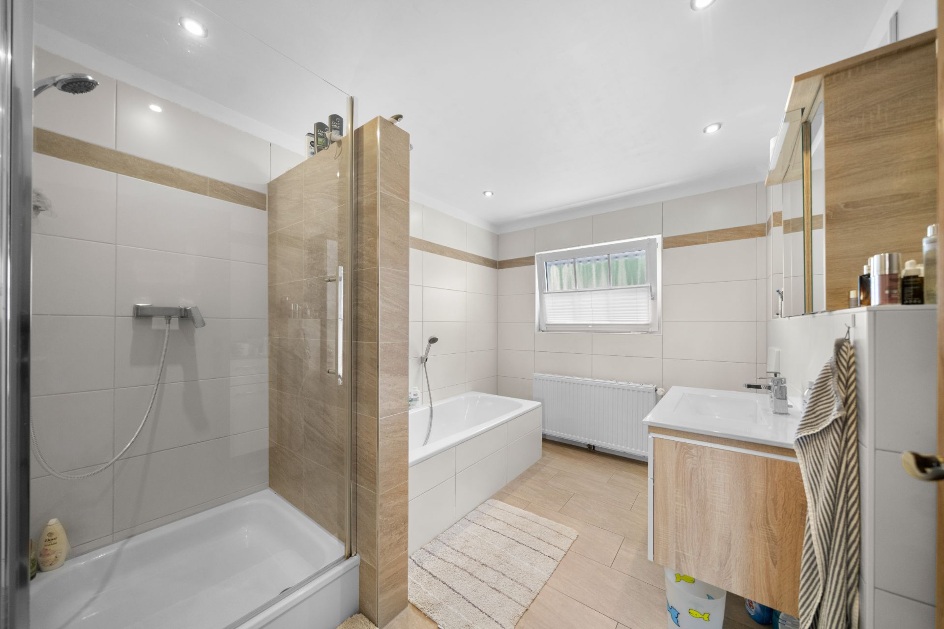 Modernes Badezimmer mit Dusche, Badewanne, Fenster und Waschbecken, helle Fliesen und Holzdetails.