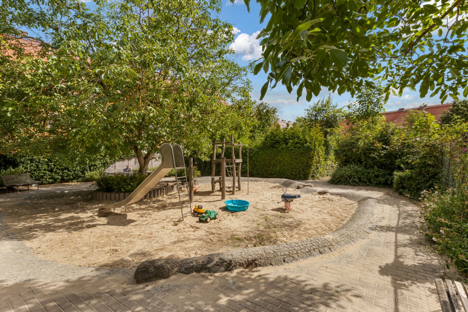 Kinderspielplatz mit Rutsche, Sandkasten und Spielzeug, umgeben von Bäumen und Hecken.