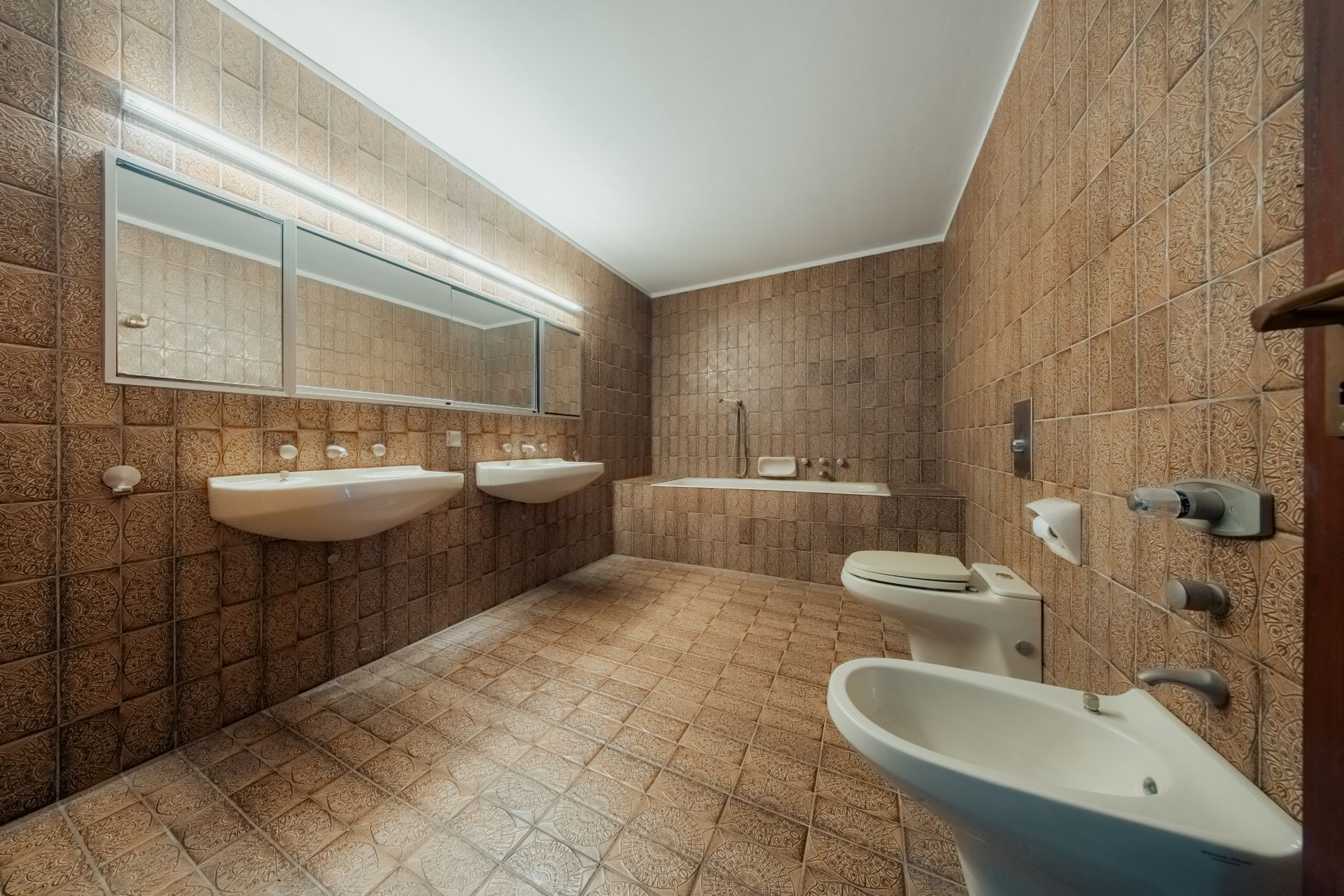 Großes Badezimmer mit braunen Fliesen, zwei Waschbecken, Spiegel, Badewanne, Toilette und Bidet.