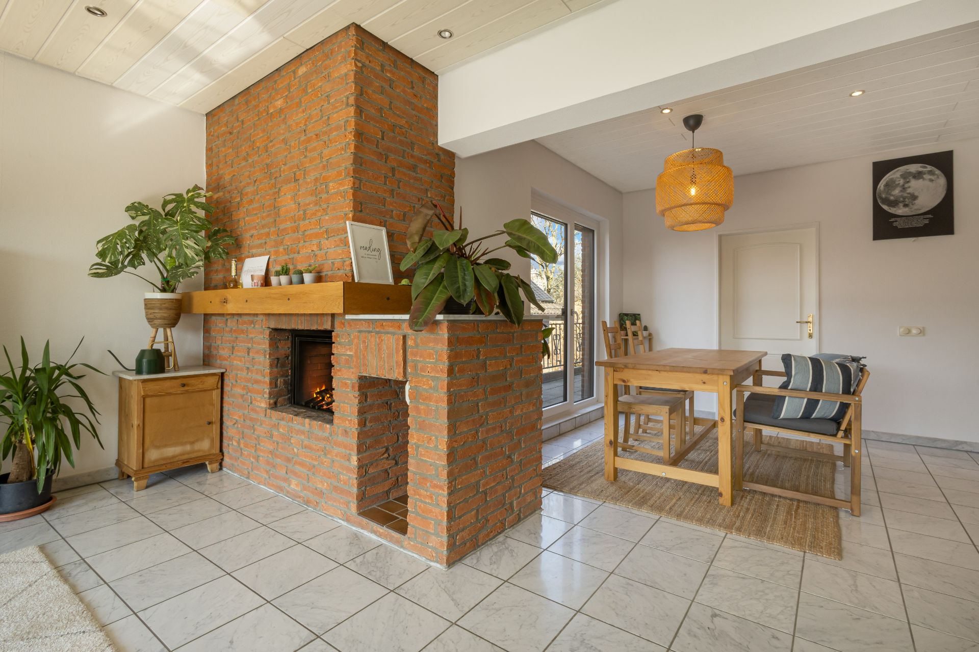 Wohnzimmer mit gemauertem Kamin, Holztisch, Pflanzen und Fliesenboden.