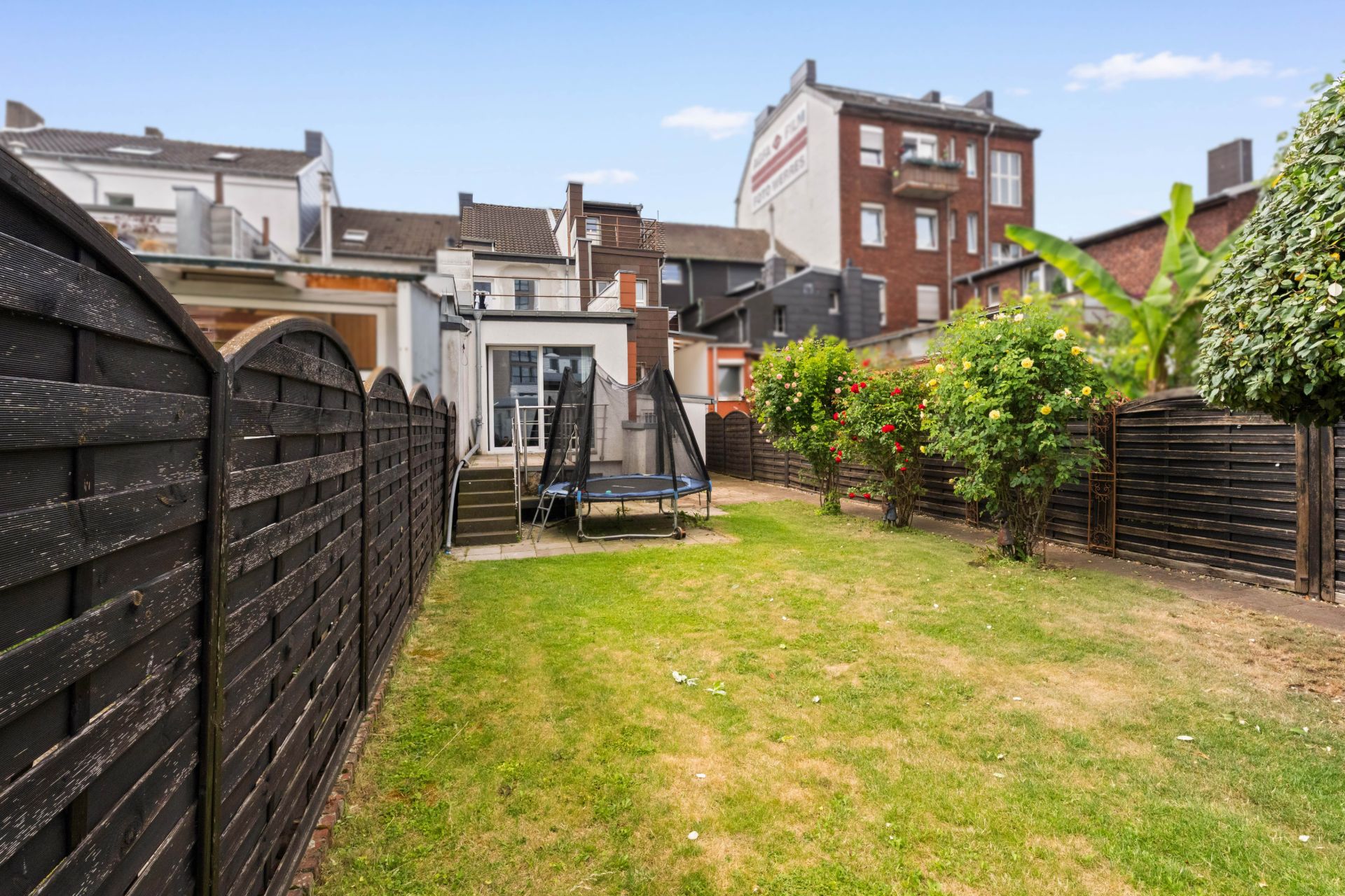 Garten mit grünem Rasen, Trampolin, Sträuchern und dunklem Holzzaun neben Wohngebäuden.