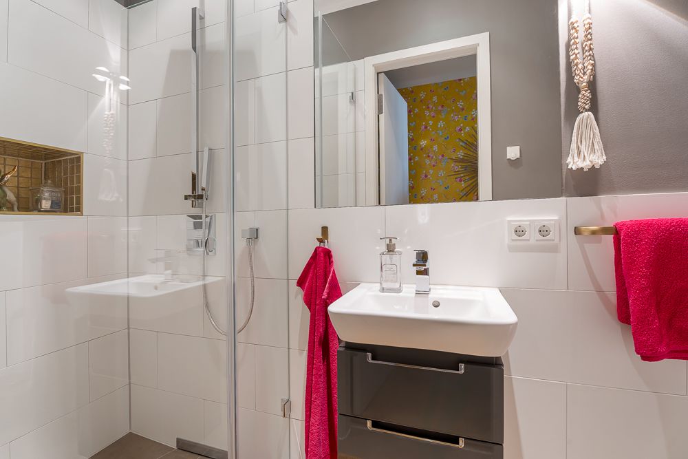 Modernes Badezimmer mit Dusche, Waschbecken und roten Handtüchern, Blick auf offene Tür mit bunter Tapete.