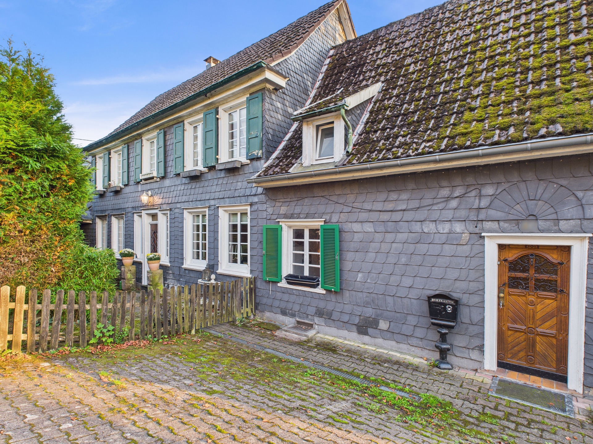 Grau gedecktes Wohnhaus mit grünen Fensterläden, Holzpforte und Zuweg aus Pflastersteinen.