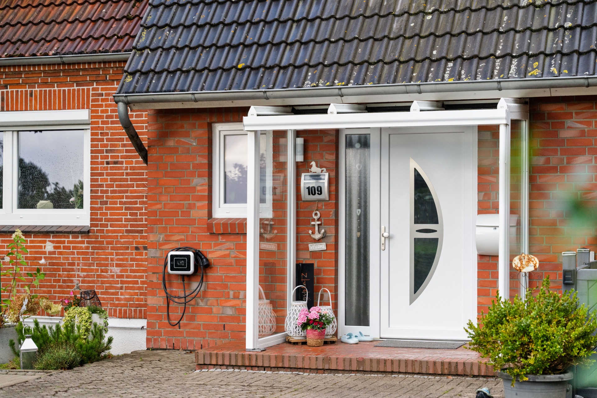 Hauseingang mit weißer Tür, überdachter Veranda und Blumen auf rotem Klinkerhaus.