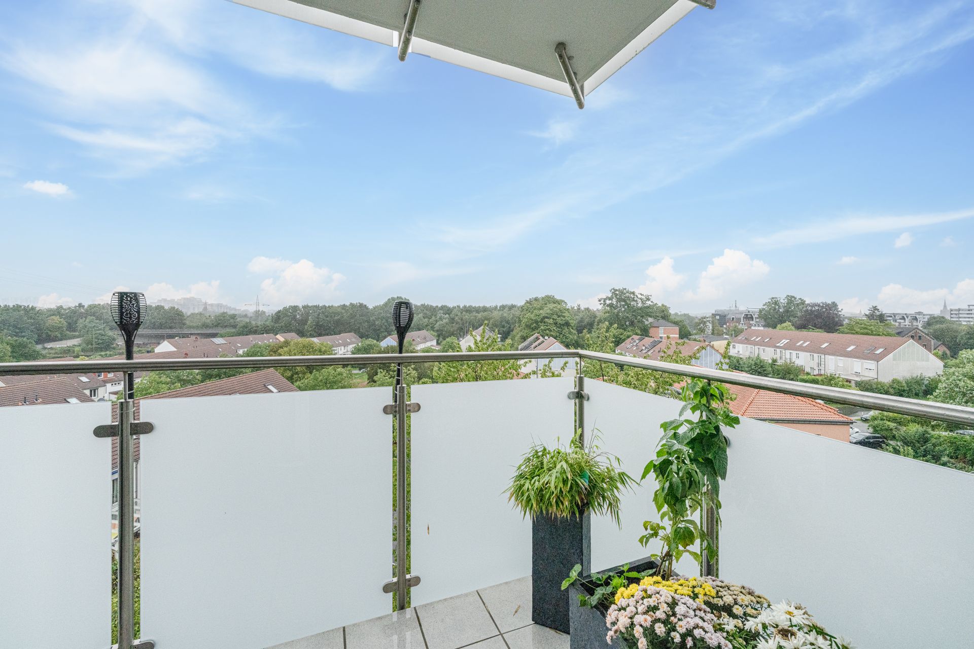 Balkon mit Pflanzenkübeln und Ausblick auf Wohnhäuser sowie Bäume unter blauem Himmel.