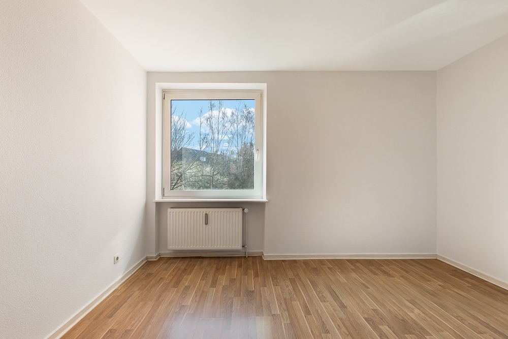 Leerer Raum mit hellem Holzboden, weißer Wand und Fenster mit Blick ins Grüne.