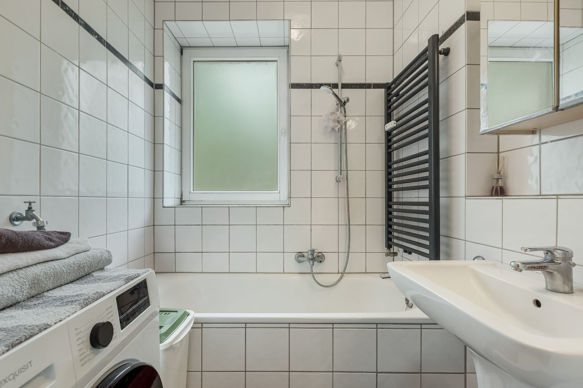 Kleines, gefliestes Bad mit Badewanne, Waschbecken, Waschmaschine und Fenster.