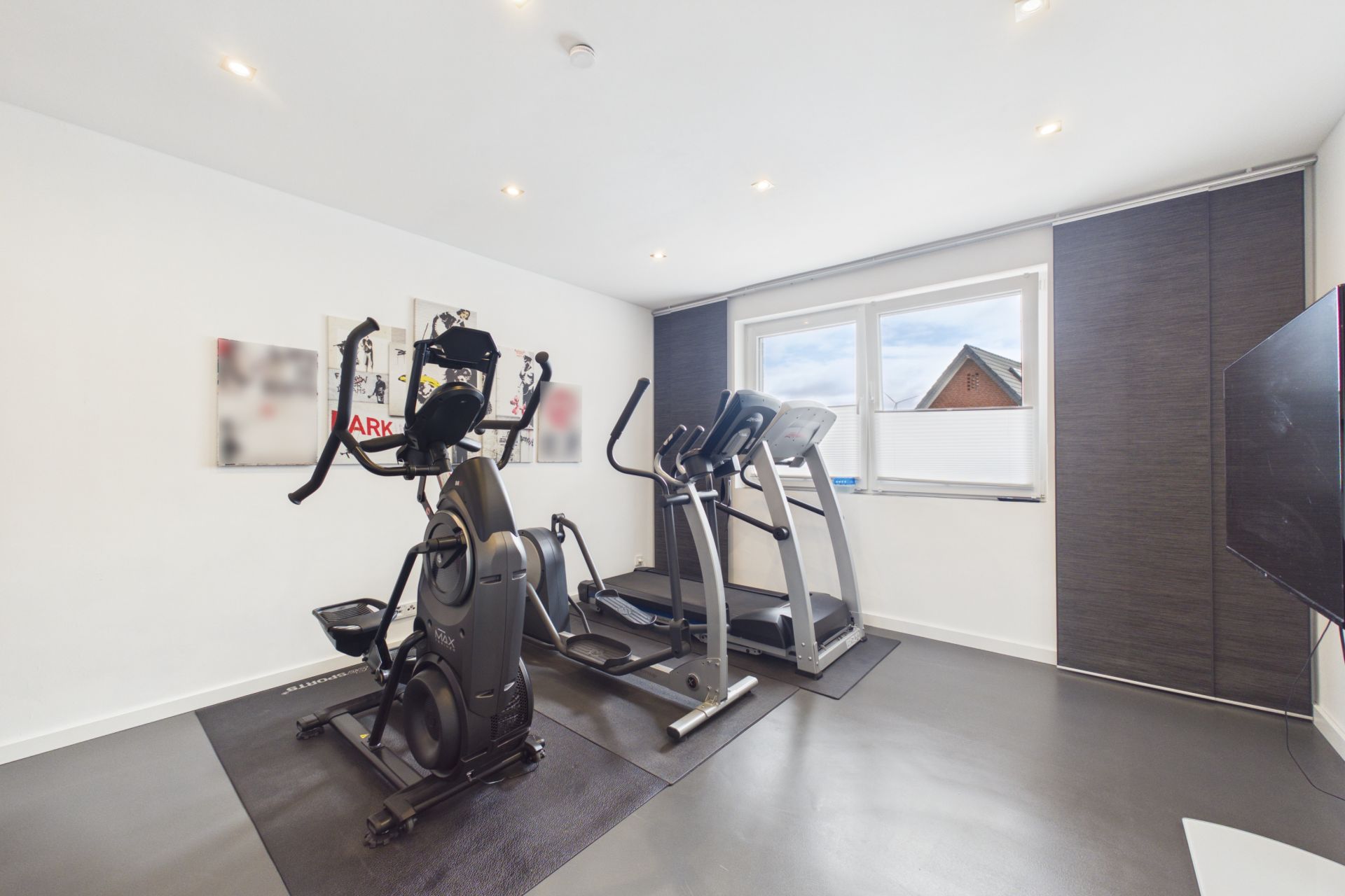Heller Raum mit zwei Fitnessgeräten, großem Fenster und Fernseher an der Wand.