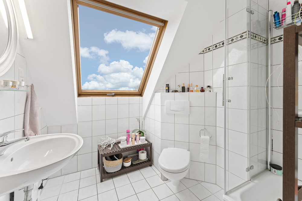 Helles Badezimmer mit Dachfenster, Dusche, Waschbecken und WC, weißen Fliesen und Regal mit Kosmetikartikeln.