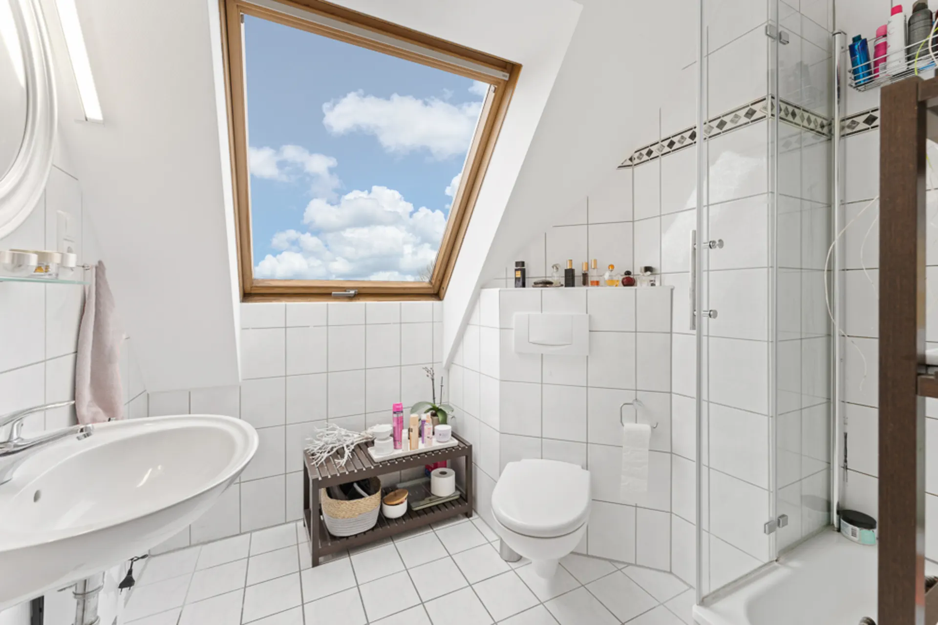 Helles Badezimmer mit Dachfenster, Dusche, Waschbecken und WC, weißen Fliesen und Regal mit Kosmetikartikeln.