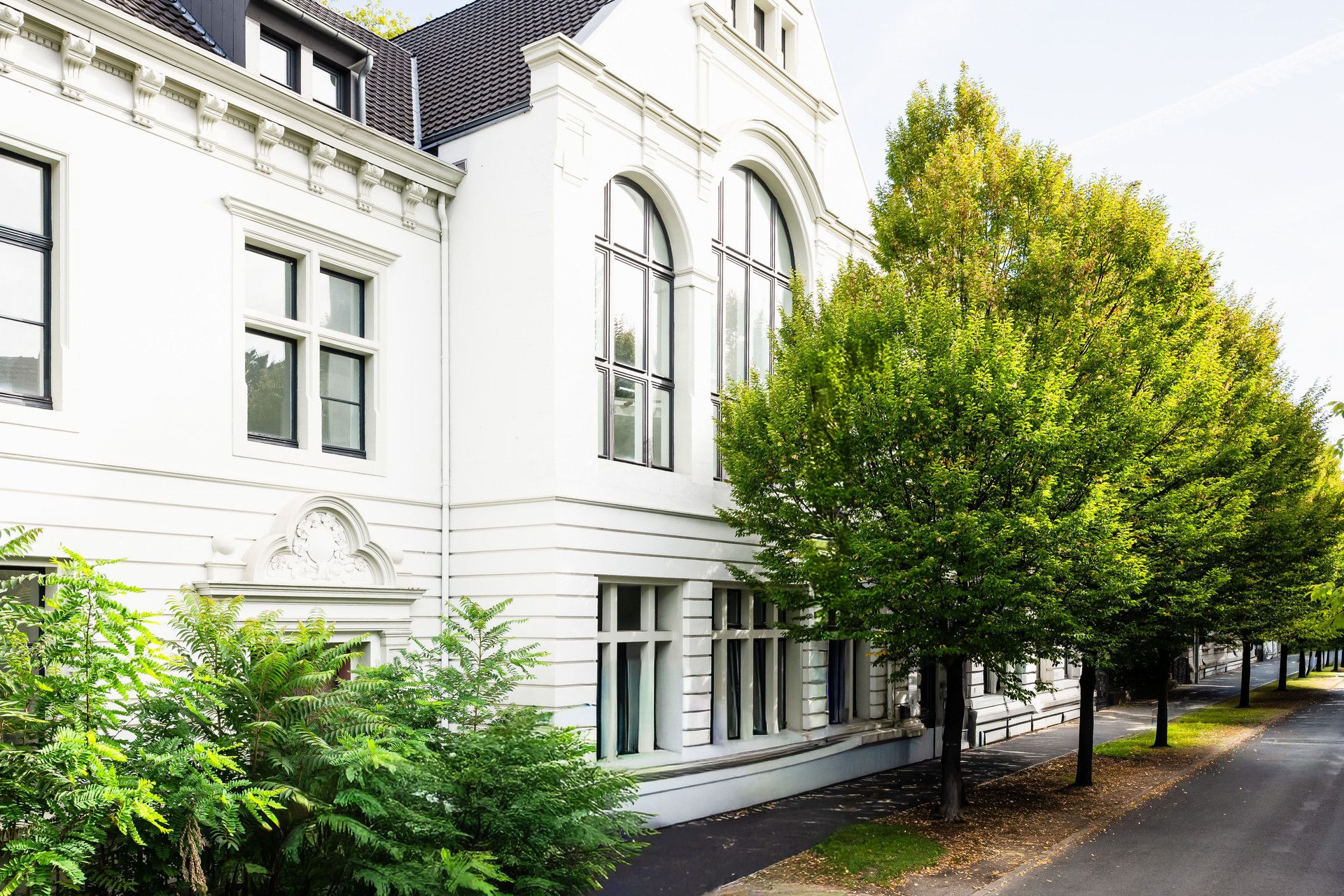 Weiße Wohnhausfassade mit großen Fenstern und Bäumen am Gehweg.