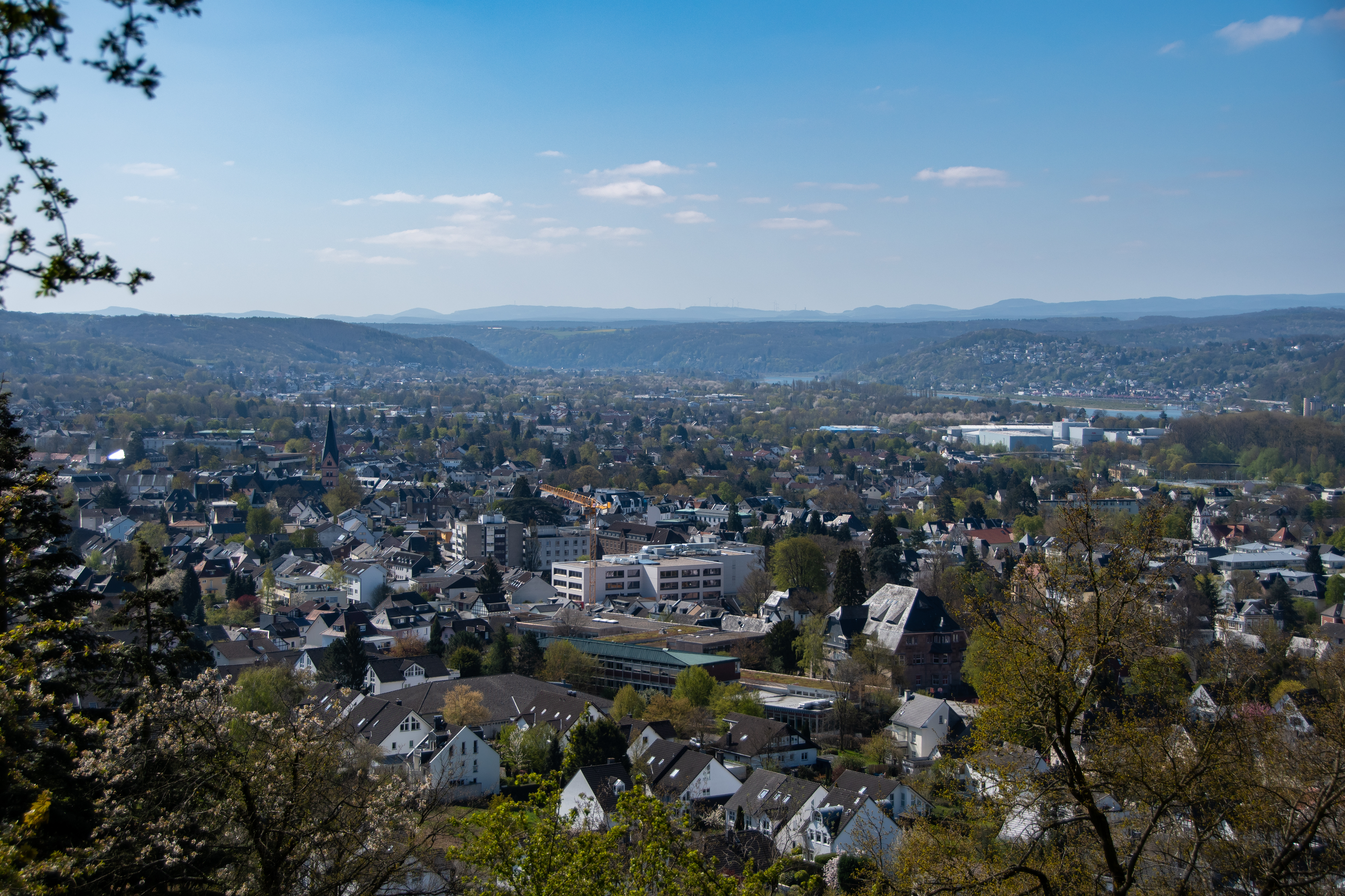 Blick über eine Wohnsiedlung mit vielen Häusern und grünen Bäumen, im Hintergrund Hügel.