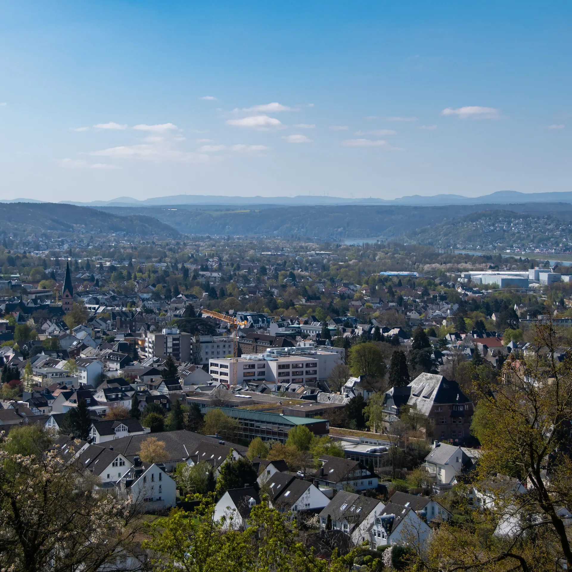 Blick über eine Wohnsiedlung mit vielen Häusern und grünen Bäumen, im Hintergrund Hügel.