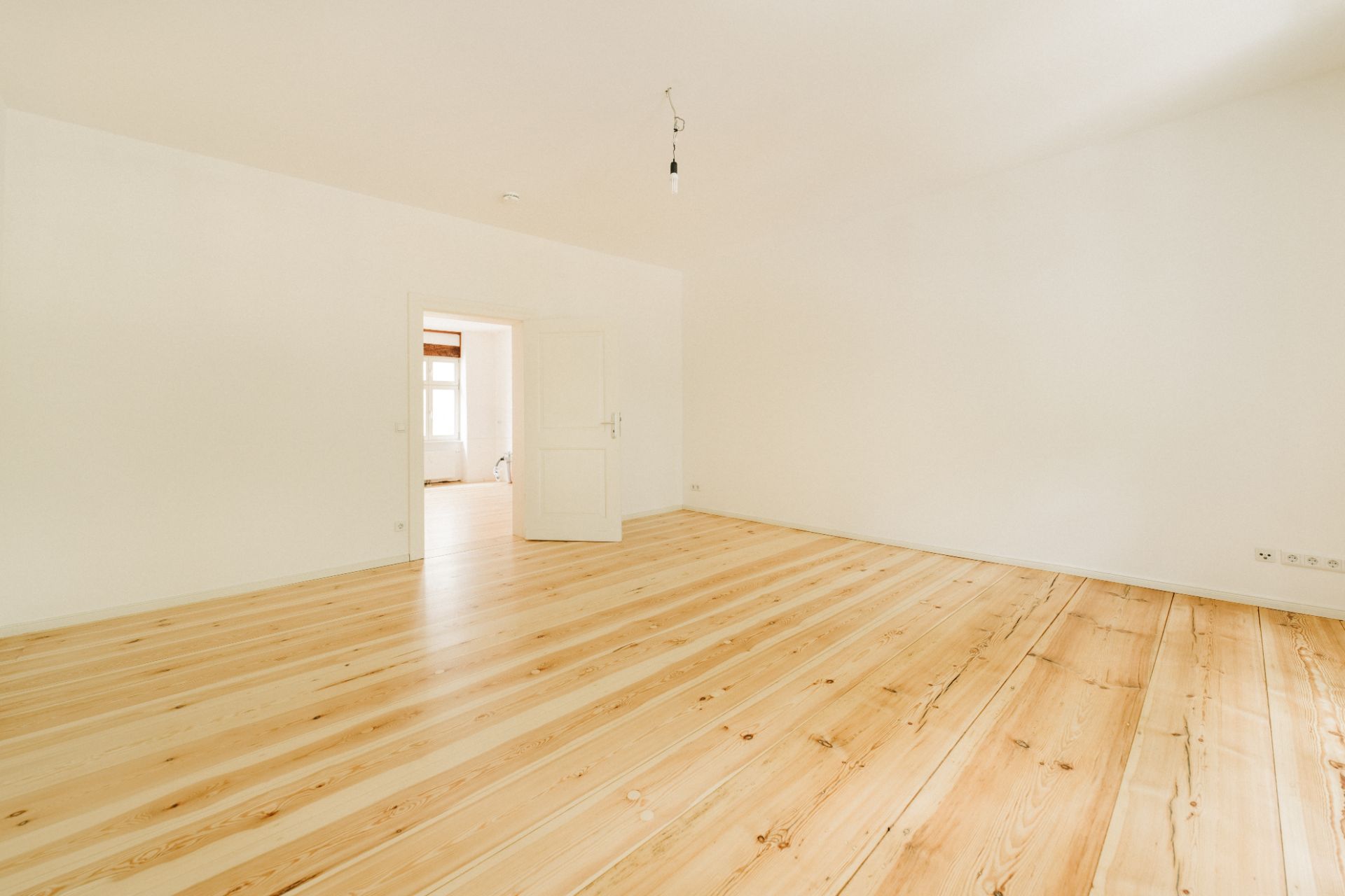 Helles, leeres Zimmer mit Holzboden und weißen Wänden, eine Tür führt in einen weiteren Raum.