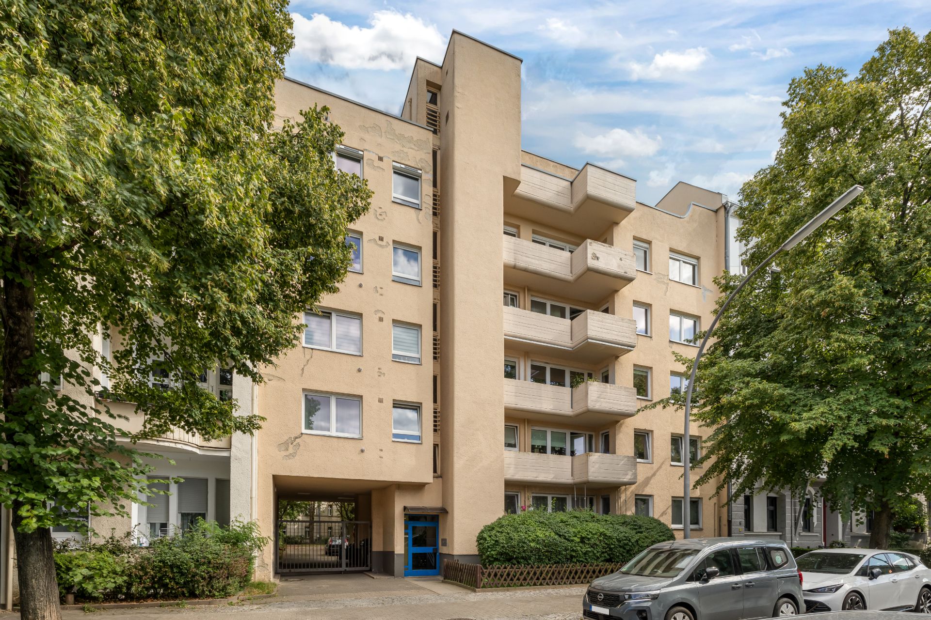 Mehrstöckiges Wohnhaus mit Balkonen und Bäumen, Autos parken auf der Straße davor.